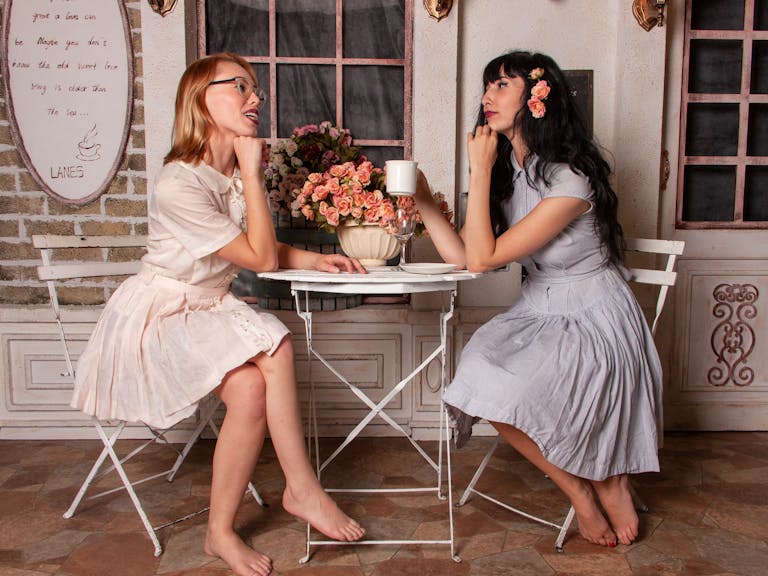 Two elegant women wearing vintage dresses enjoying conversation and coffee at a cozy indoor café setting.